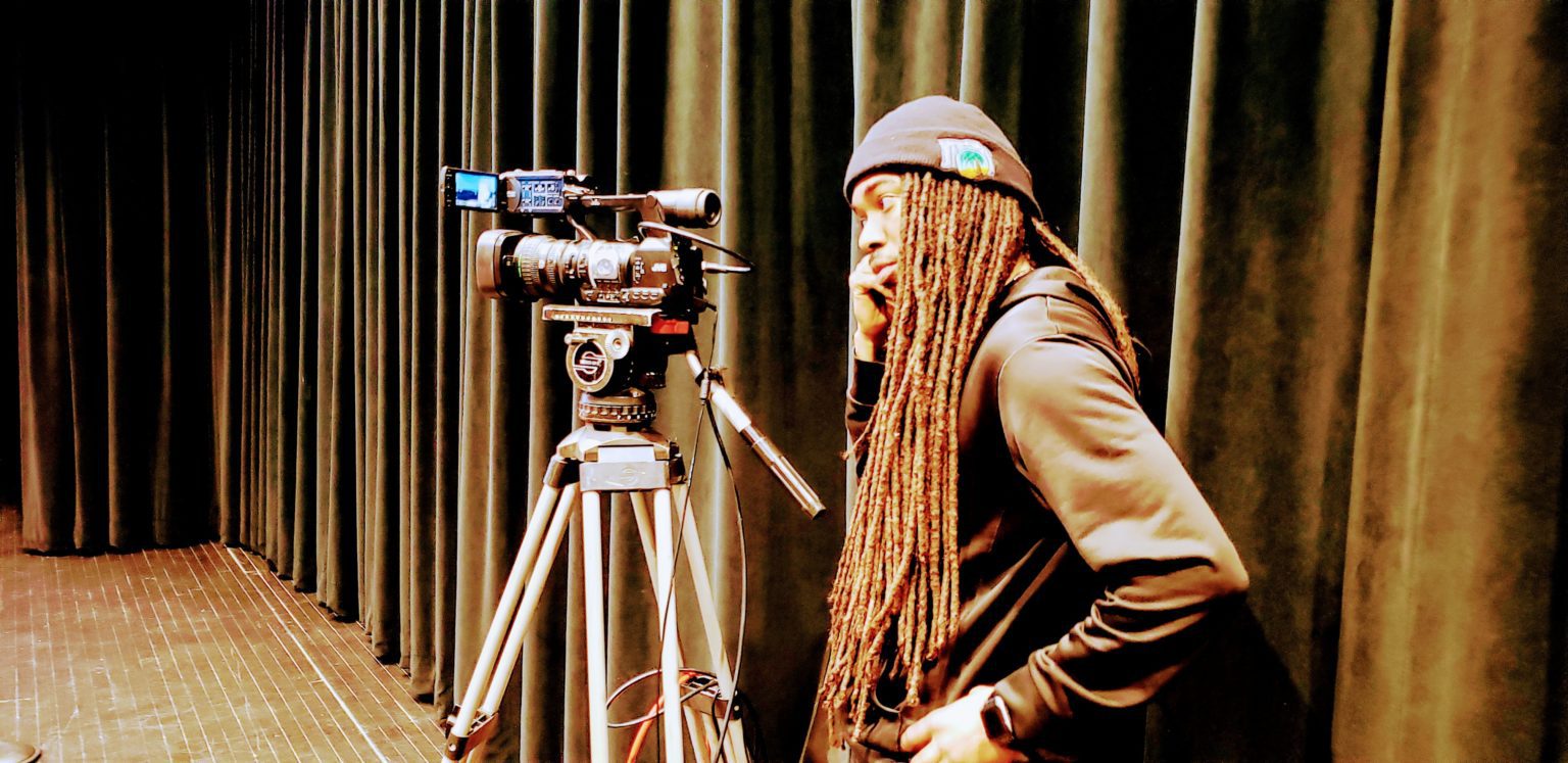 A man with dreadlocks standing in front of a camera.
