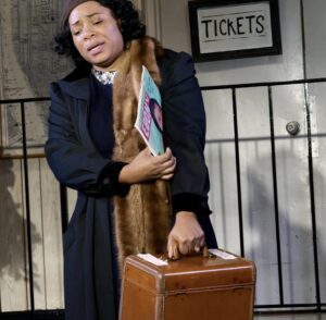 A woman in a fur coat and scarf holding onto an old suitcase.